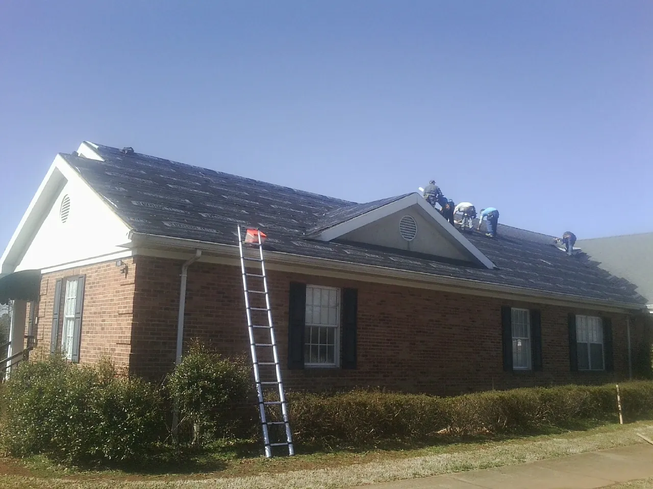 Crew tearing shingles off a brick ranch roof with a ladder set — Metal Roof Repair work by our crew in Herscher