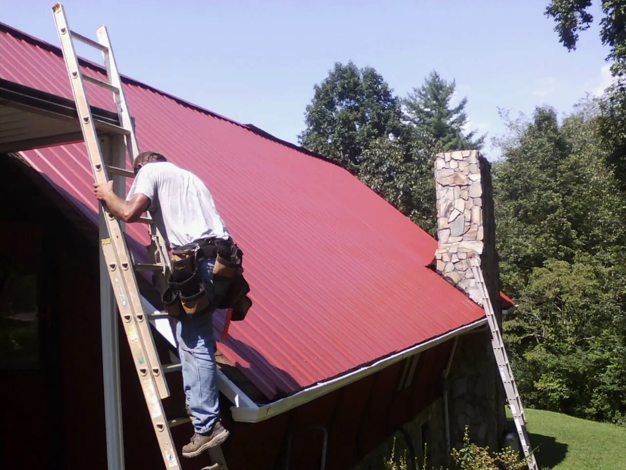 Roofer on a ladder beside a red metal roof and stone chimney — Soffit & Fascia Repair by Roofing Brothers in Herscher